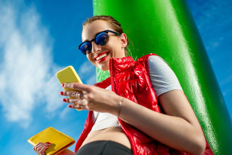 Mujer con chaleco rojo acolchado y gafas de sol sonriendo mientras usa un smartphone amarillo al aire libre, sosteniendo una libreta amarilla a juego, contra un fondo vibrante azul y verde